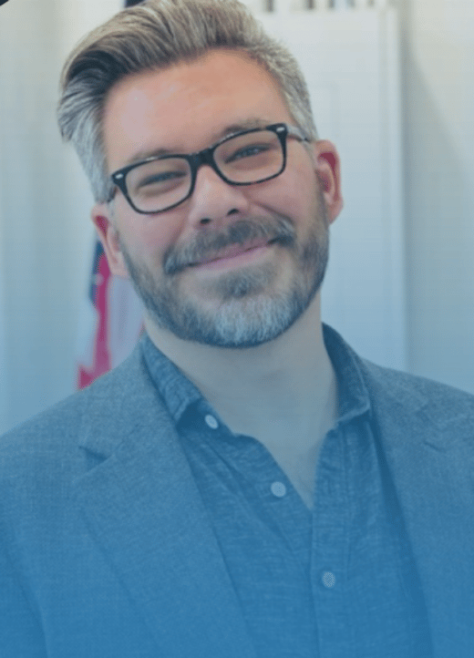 Danny Combs smiles at the camera, wearing glasses and a blazer.